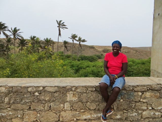 Farbige Frau in rotem Shirt sitzt auf einer Mauer vor einer grünen Landschaft auf Maio, Kapverden