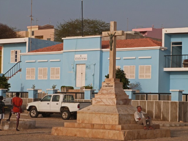 Hellblaues Haus, davor ein Monument mit Jesus am Kreuz und Mann, der darunter sitzt, Maio, Kapverden