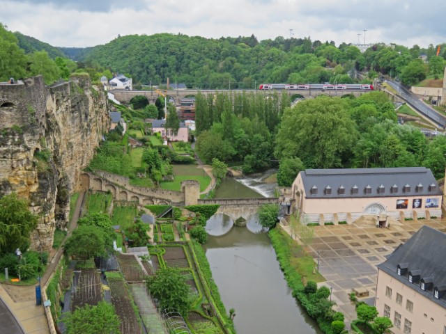 Blick über Luxemburg Stadt mit der Stadtmauer, Häusern, einen Wassergraben und einer Brücke mit Zug darauf