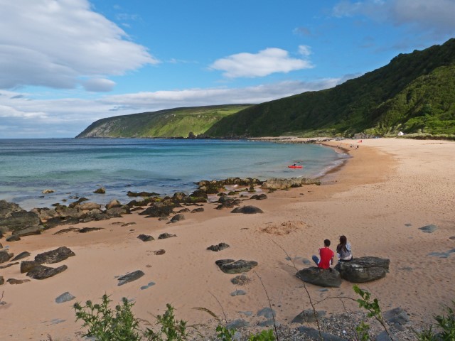 Zwei Leute sitzen auf einem Stein an einem Sandstrand und schauen auf das Meer und grüne Felsen, Donegal, Irland