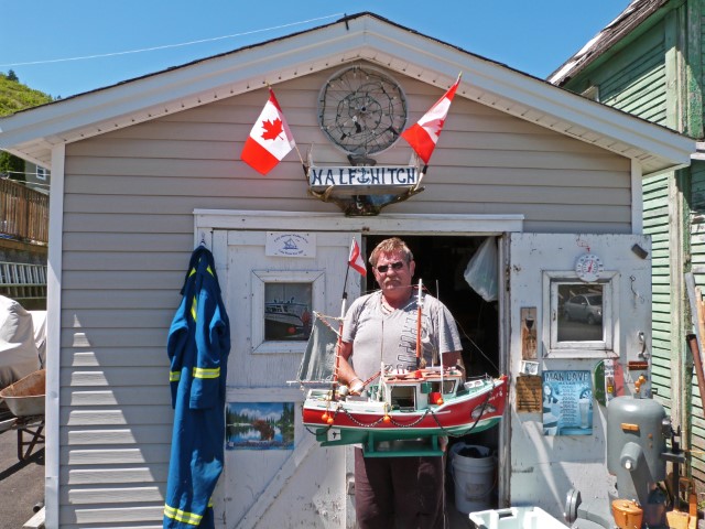 Mann mit Holzbootmodell in den Händen vor einer Hütte mit kanadischen Flaggen, Neufundland