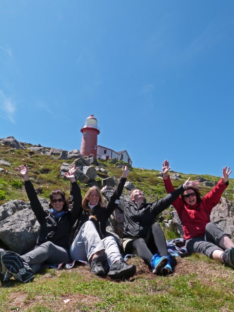 Vier Frauen sitzen auf Picknickdecken unterhallb des Ferryland Lighthouses und werfen begeistert die Arme in die Luft, Neufundland