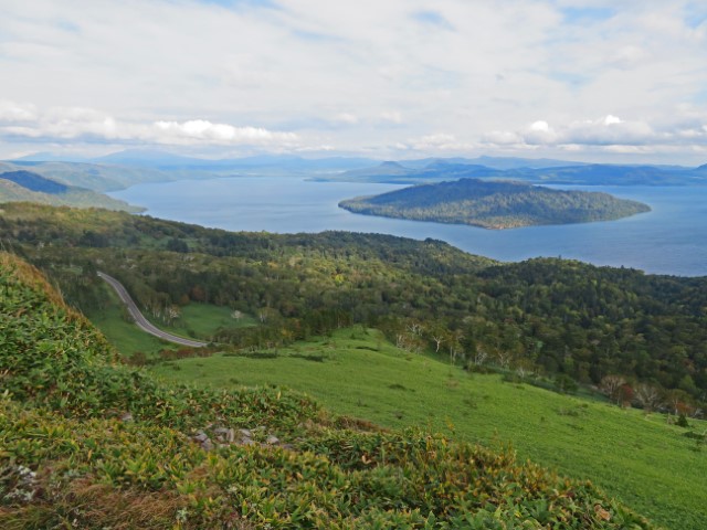 Blick über den Kussharo See mit einer Insel in der Mitte und umgeben von viel Grün, Hokkaido, Japan