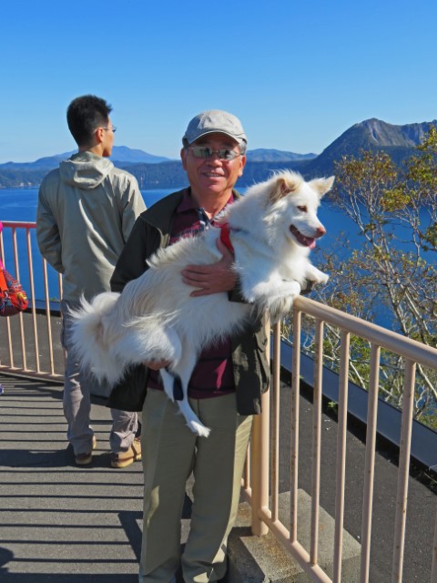 Japaner mit Sonnenbrille und großem weißem Hundeim Arm am Aussichtspunkt über den Mashu See