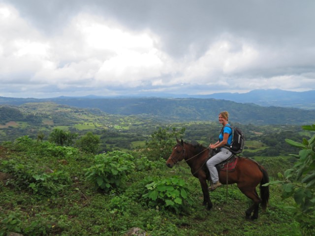 Junge Frau auf braunem Pferd vor grüner hügeliger Landschaft in Nicaragua