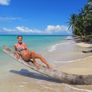 Heiße junge Frau in Bikini auf einem umgefallenen Baumstamm am weißen Strand mit blauem Meer auf Little Corn Island