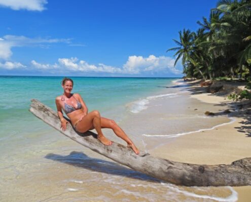 Heiße junge Frau in Bikini auf einem umgefallenen Baumstamm am weißen Strand mit blauem Meer auf Little Corn Island