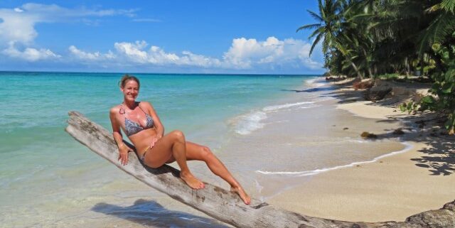 Heiße junge Frau in Bikini auf einem umgefallenen Baumstamm am weißen Strand mit blauem Meer auf Little Corn Island