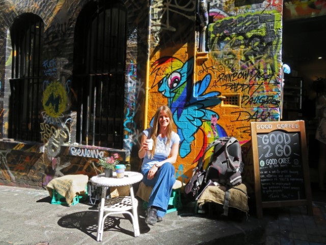 Junge Frau sitzt mit Kaffeebecher vor einer mit Street Art bemalten Wand in einem Café in Melbourne