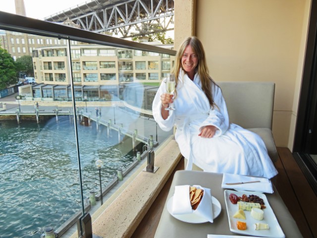 Blonde Frau in Bademantel sitzt auf Balkon des Park Hyatt Sydney mit einem Glas Champagner in der Hand