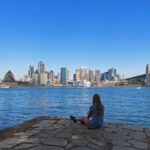 Frau sitzt am Wasser mit Blick übers Wasser und die Skyline von Sydney mit dem Opernhaus