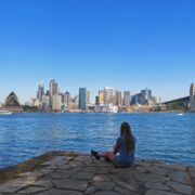 Frau sitzt am Wasser mit Blick übers Wasser und die Skyline von Sydney mit dem Opernhaus