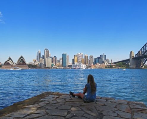 Frau sitzt am Wasser mit Blick übers Wasser und die Skyline von Sydney mit dem Opernhaus