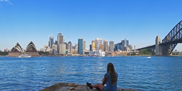 Frau sitzt am Wasser mit Blick übers Wasser und die Skyline von Sydney mit dem Opernhaus