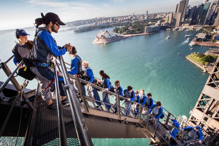 Menschen laufen eine steile Treppe hinauf auf die Sydney Harbour Bridge mit Panorama von Hafen und Opernhaus