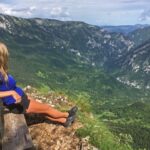 Junge Frau in Shorts sitzt auf einer Bank auf einem Gipfel und schaut über die Berge Montenegros