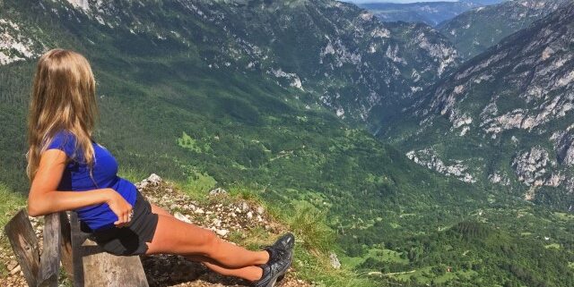 Junge Frau in Shorts sitzt auf einer Bank auf einem Gipfel und schaut über die Berge Montenegros