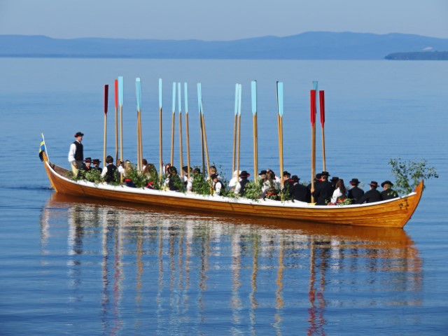 Traditionelles Kirchenboot auf dem Siljansee mit traditionell gekleideten Leuten, die ihre Rude in die Höhe halten