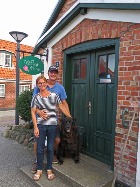 Ehepaar vor einem Klinkerhaus, daran ein Schild "Wedding Island"