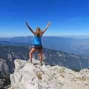 Junge Wanderfrau auf einem Fels blickt über Berglandschaft und wirft die Arme in die Höhe