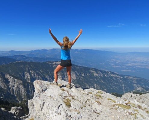 Junge Wanderfrau auf einem Fels blickt über Berglandschaft und wirft die Arme in die Höhe