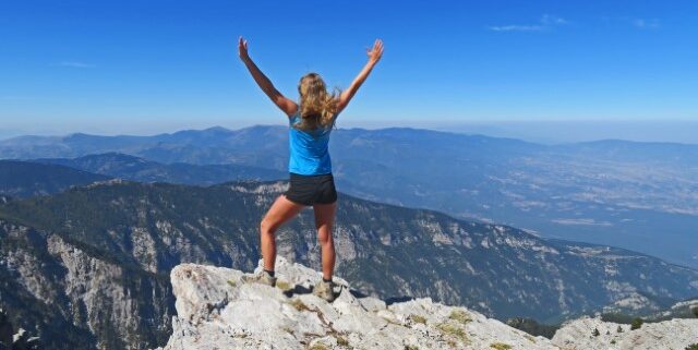 Junge Wanderfrau auf einem Fels blickt über Berglandschaft und wirft die Arme in die Höhe