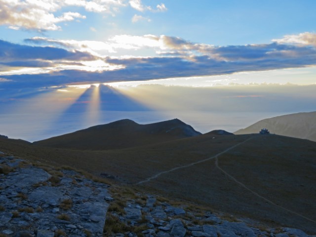 Die Sonbne bricht durch Wolken über dem Meer, im Vordergrund die Berge