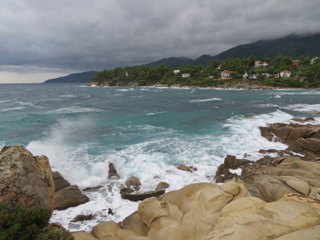 Blick über helle Felsen, an denen das blaue Meer hochtost, im Hintergrund begrünte Klippen