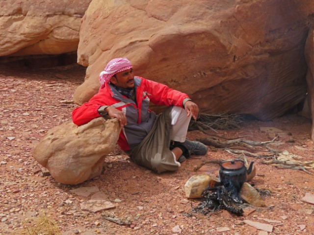 Beduine mit roter Jacke und Kopftuch kocht or einem Felsen über dem Feuer Tee