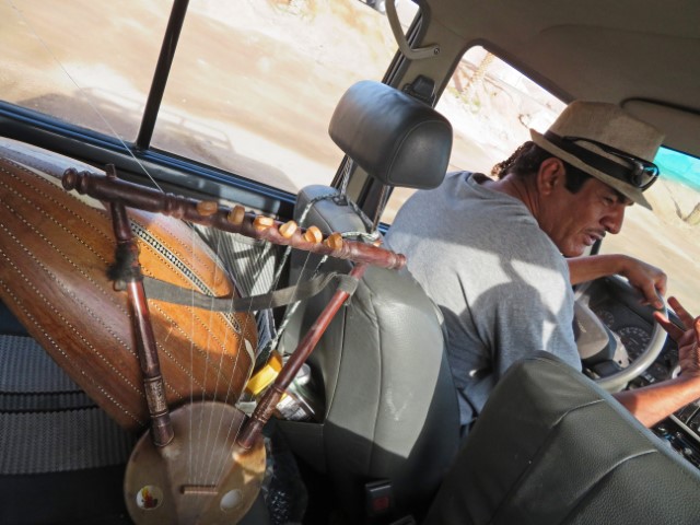 Mann mit Hut fährt Auto, auf dem Nebensitz liegt ein Musikinstrument mit Saiten namens Oud