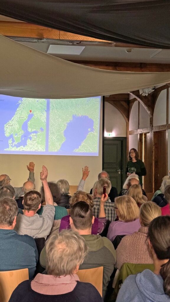 Zuschauer heben die Hand beim Multimedia-Vortrag von Bernadette Olderdissen, auf der Leinwand eine Karte Schwedens